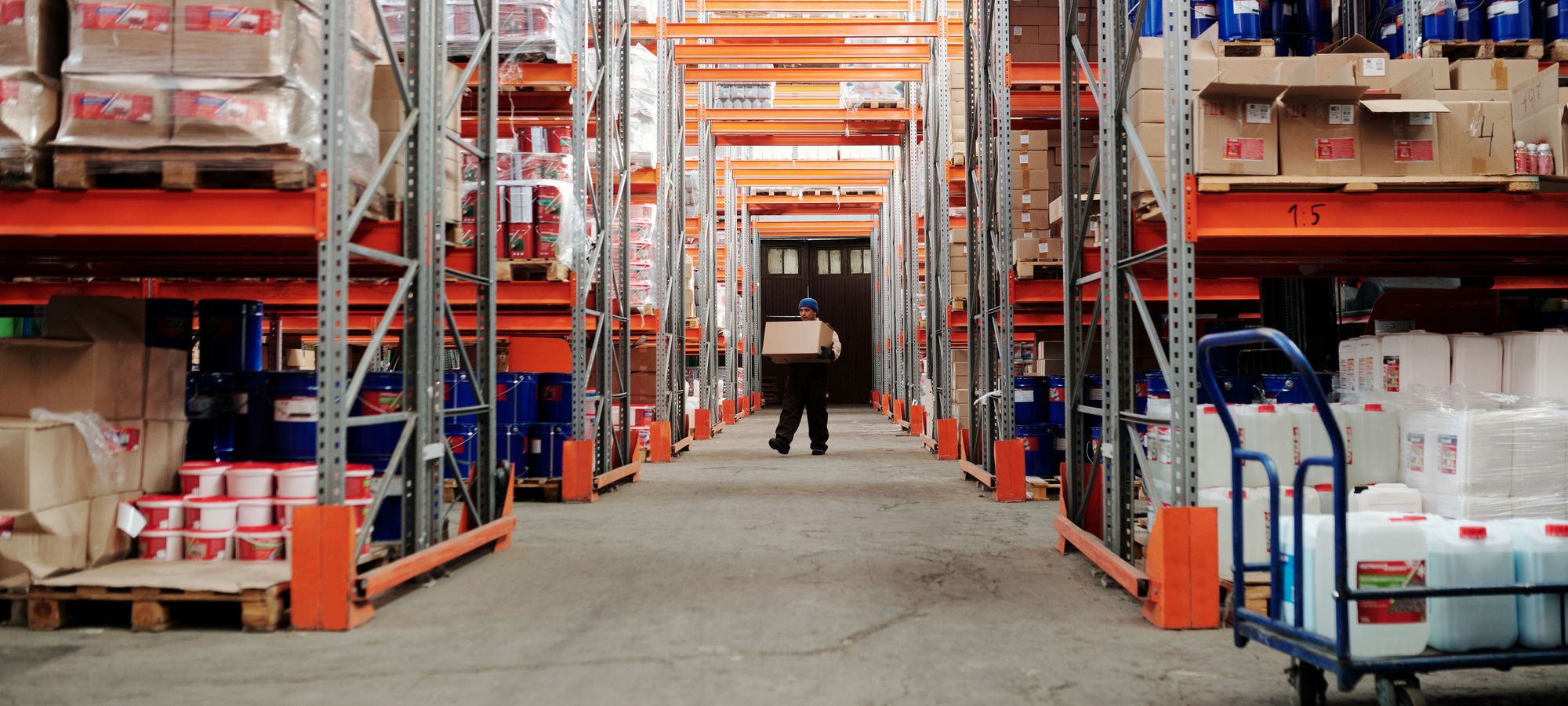 A worker carrying a box down an aisle of racked warehouse shelving stocked with cases and containers